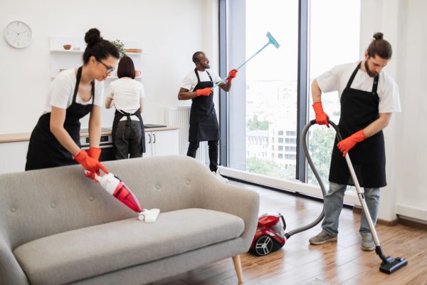 Professional team of people of cleaning service cleans sofa, carpet, cuisine table and panoramic windows of spacious apartment. Multinational group of cleaners cleans bright studio kitchen.
